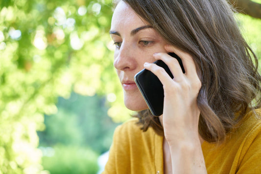 Side profile of a woman using a smartphone outdoors in a sunlit park, wearing a yellow top.