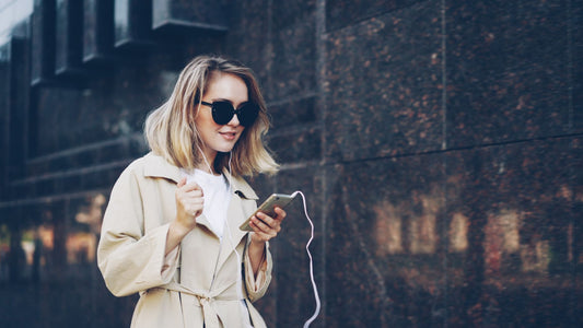 Fashionable woman with sunglasses enjoys music on smartphone outdoors.