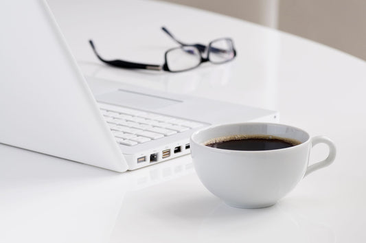 A minimalist workspace featuring a laptop, a cup of black coffee, and eyeglasses on a white desk.