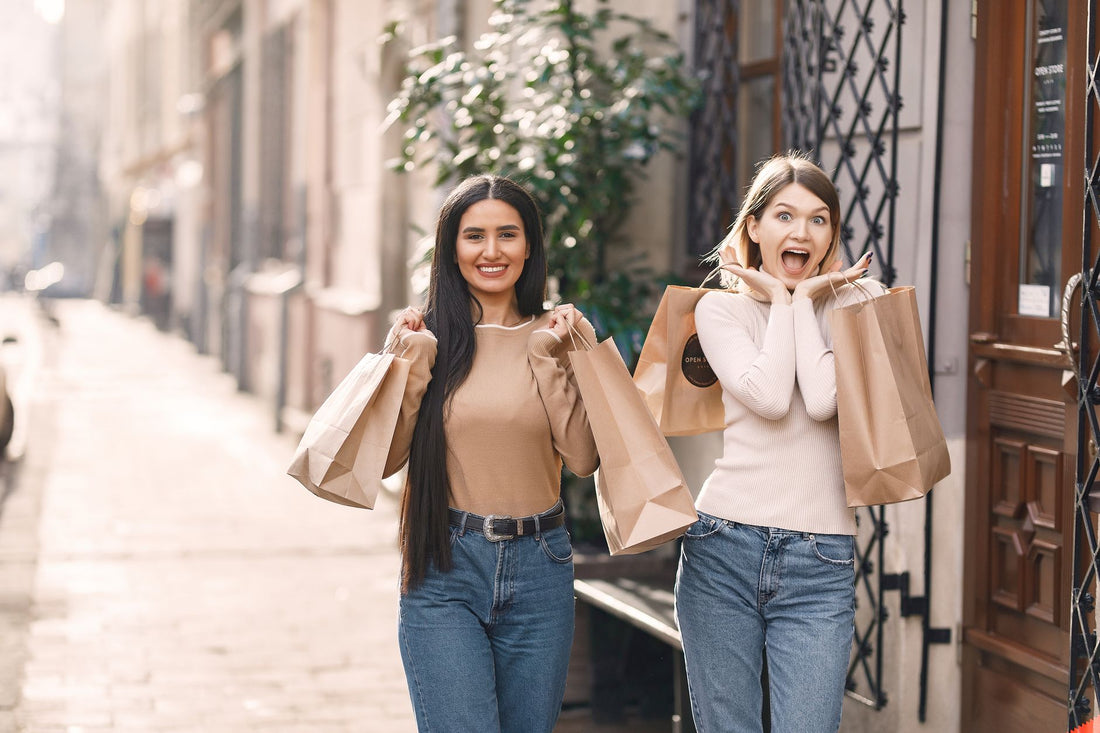 Positive young casually dressed women with shopping bags standing near building while smiling and looking at camera in sunny summer day