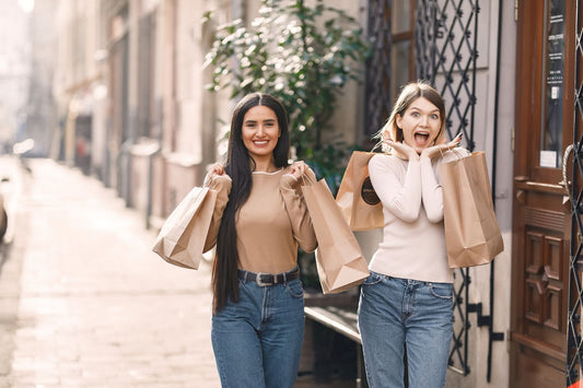 Positive young casually dressed women with shopping bags standing near building while smiling and looking at camera in sunny summer day