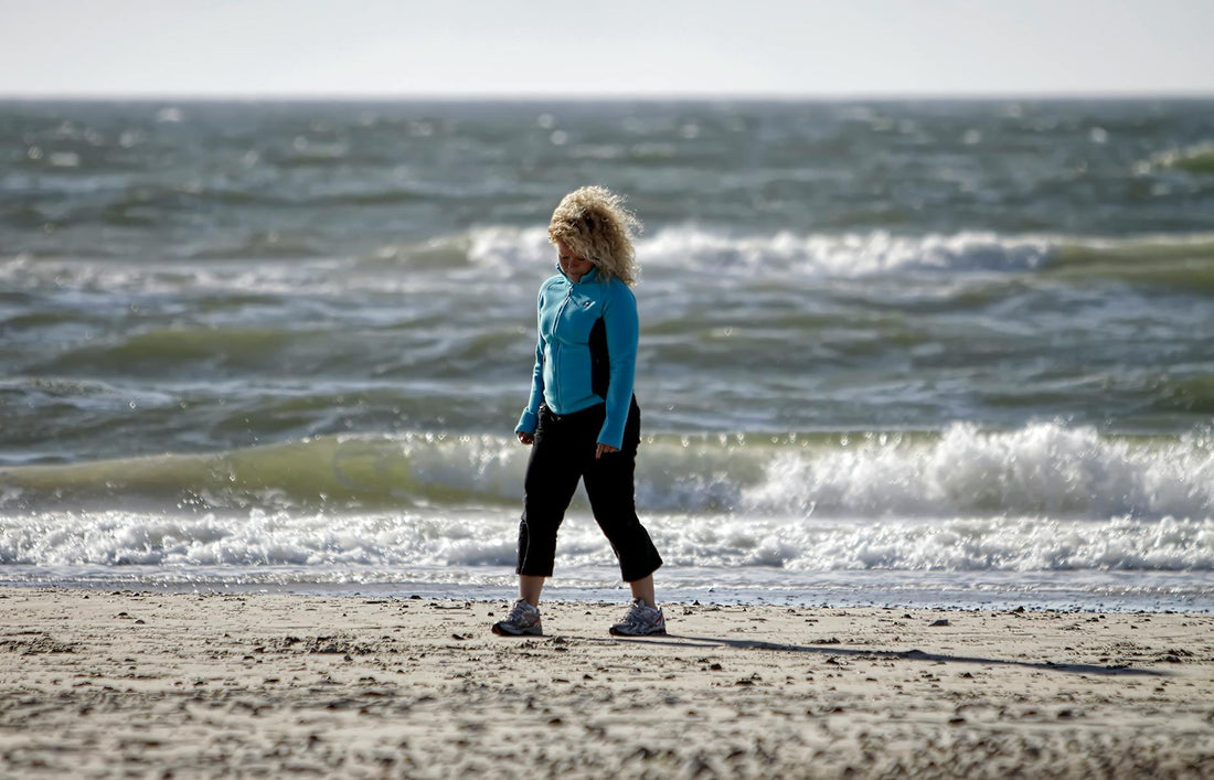 A woman in activewear walks along a windy beach in Denmark, with ocean waves crashing behind her.
