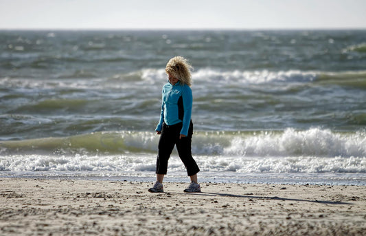 A woman in activewear walks along a windy beach in Denmark, with ocean waves crashing behind her.