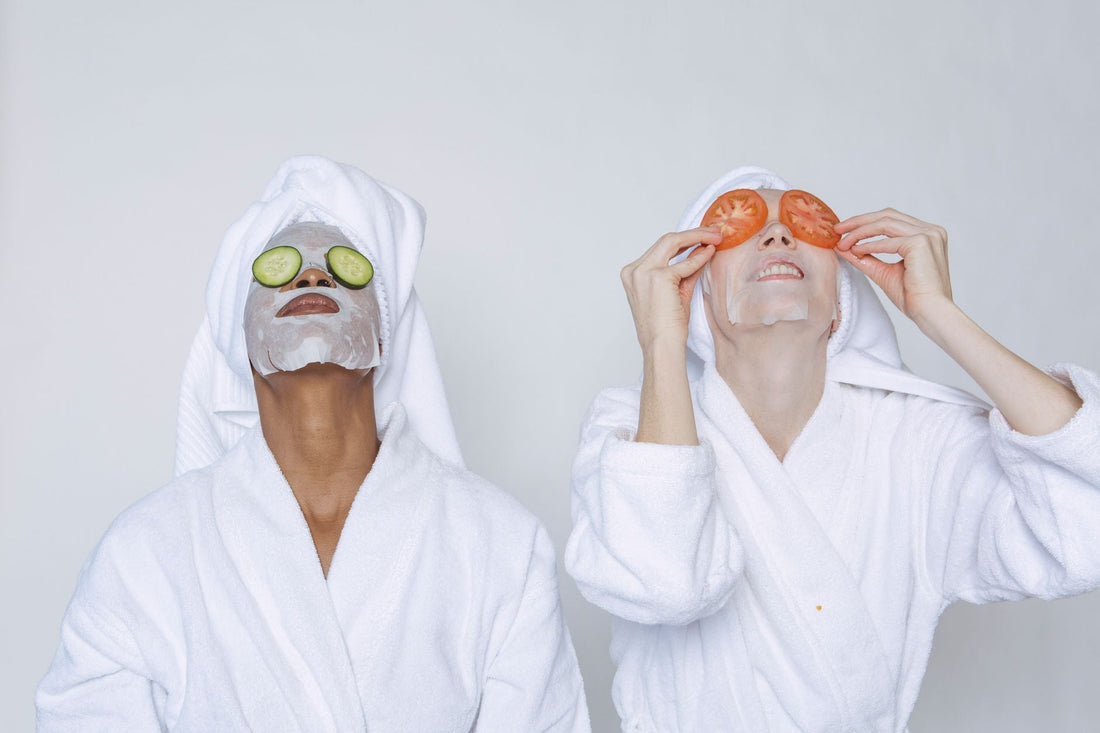 Young multiethnic females wearing bathrobes and towels moisturizing face with masks and slices of tomatoes and cucumbers against white background